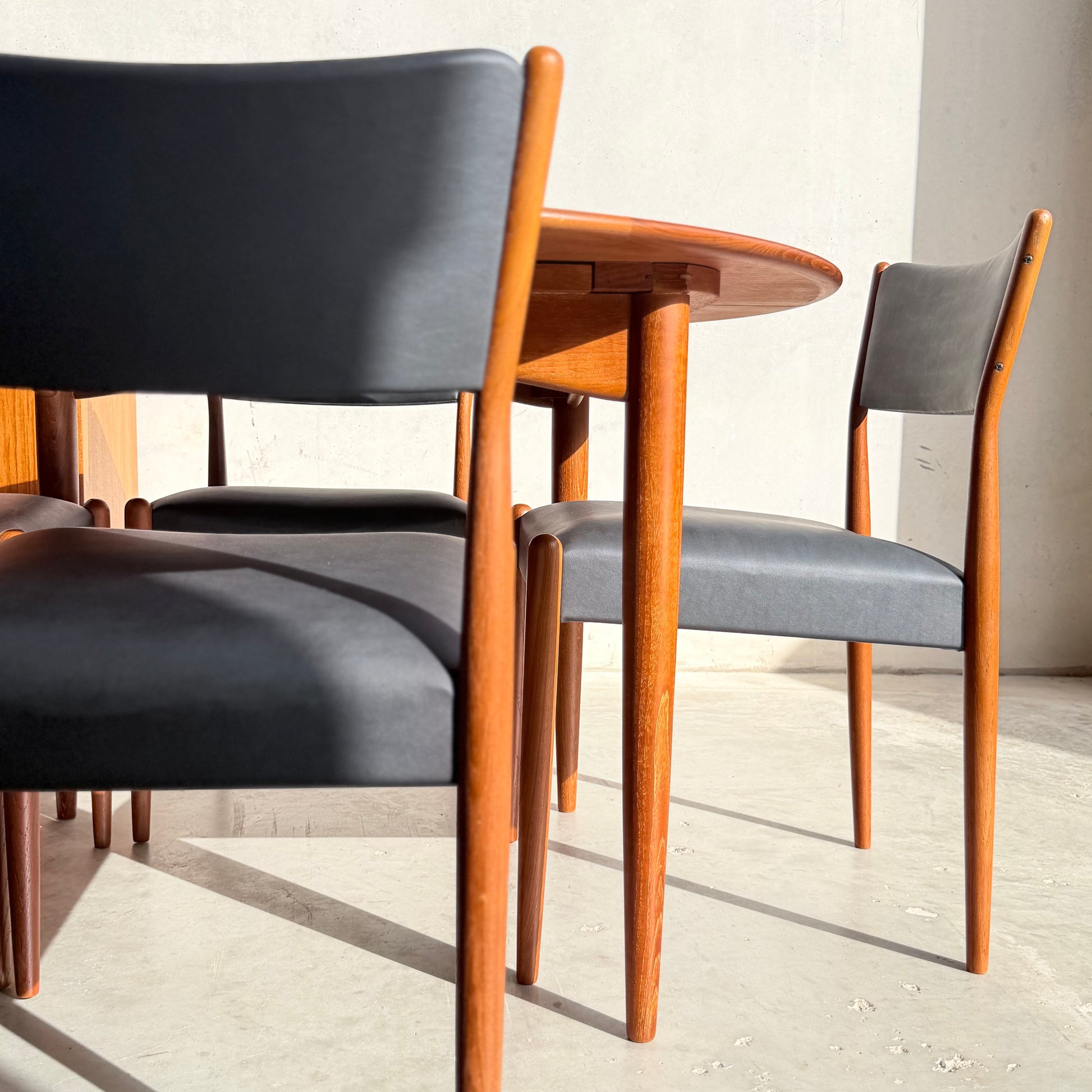 Sunlight highlights a set of Parker Number 97 Dining Chairs Set of 4 – Licorice Vinyl (Rediscover) around a round wooden table, casting graceful shadows on the light floor and wall.