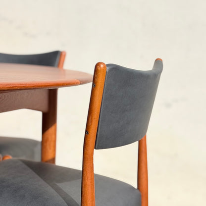 A close-up of Parker Number 97 Dining Chairs Set of 4 – Licorice Vinyl (Rediscover) shows a dark backrest beside a round wooden table, both casting soft shadows against a light background.