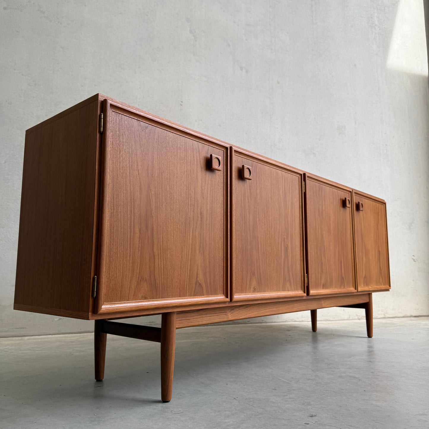 The Iconic Parker Teak 4-Door Sideboard – Restored Mid Century Modern with Tapered Legs (Rediscover) features square handles and stands against a plain grey wall and concrete floor, bathed in soft natural light.