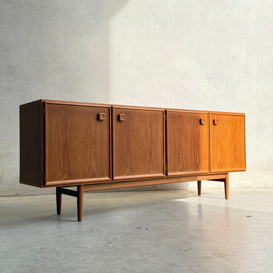 The Iconic Parker Teak 4-Door Sideboard – Restored Mid Century Modern with Tapered Legs (Rediscover) displays refined Australian craftsmanship, four cabinet doors, and sleek tapered legs against a softly sunlit light gray wall.