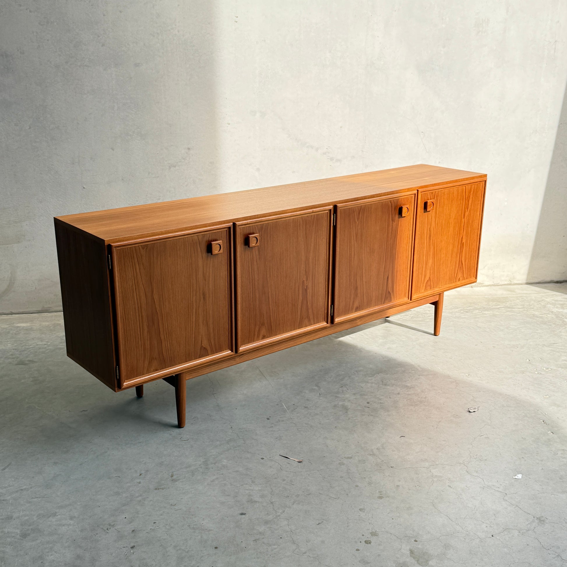 The Iconic Parker Teak 4-Door Sideboard—restored mid-century modern with tapered legs—exemplifies Australian craftsmanship, featuring classic design and sleek lines, displayed against a light gray wall on a concrete floor.