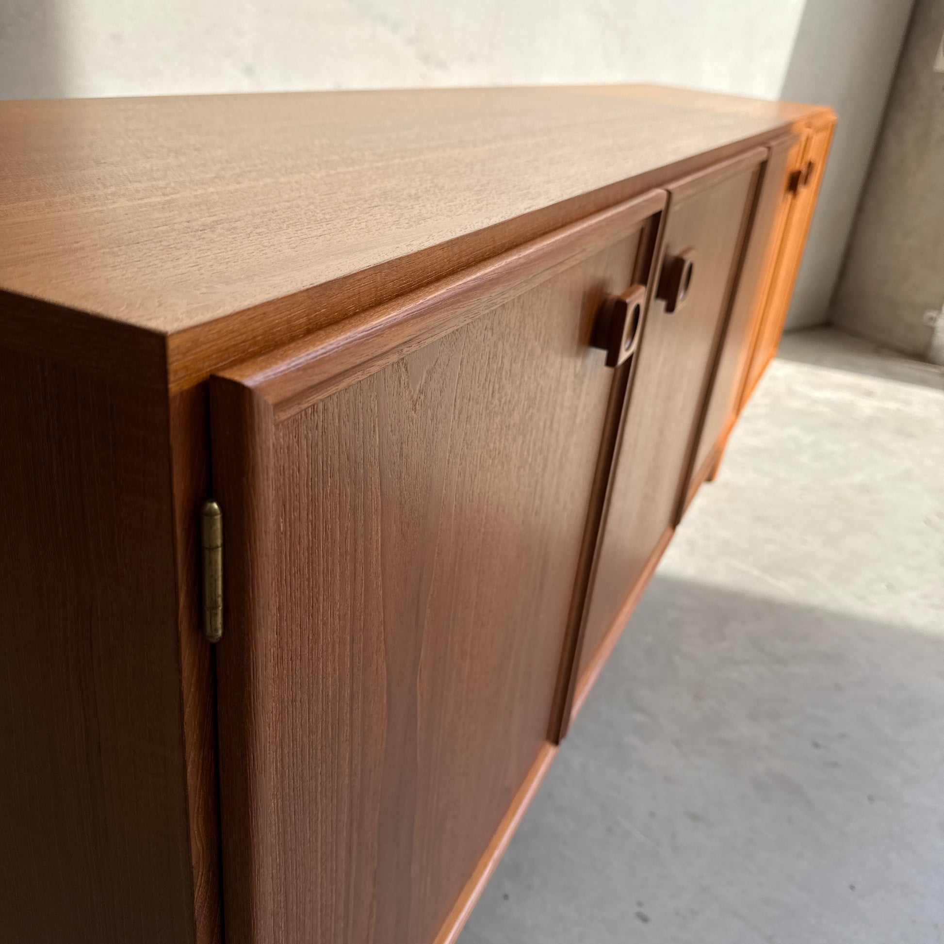 A close-up of the Iconic Parker Teak 4-Door Sideboard shows its cabinet doors, square handles, and brass hinges on a concrete floor in sunlight—a restored mid-century modern piece with tapered legs from renowned Australian craftsmanship.