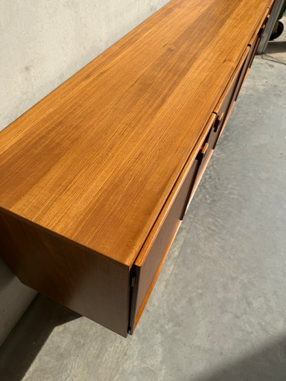 The Iconic Parker Teak 4-Door Sideboard – Restored Mid Century Modern with Tapered Legs (Rediscover) sits against a light wall on a concrete floor, its smooth finish and Australian craftsmanship visible from an angled top view.