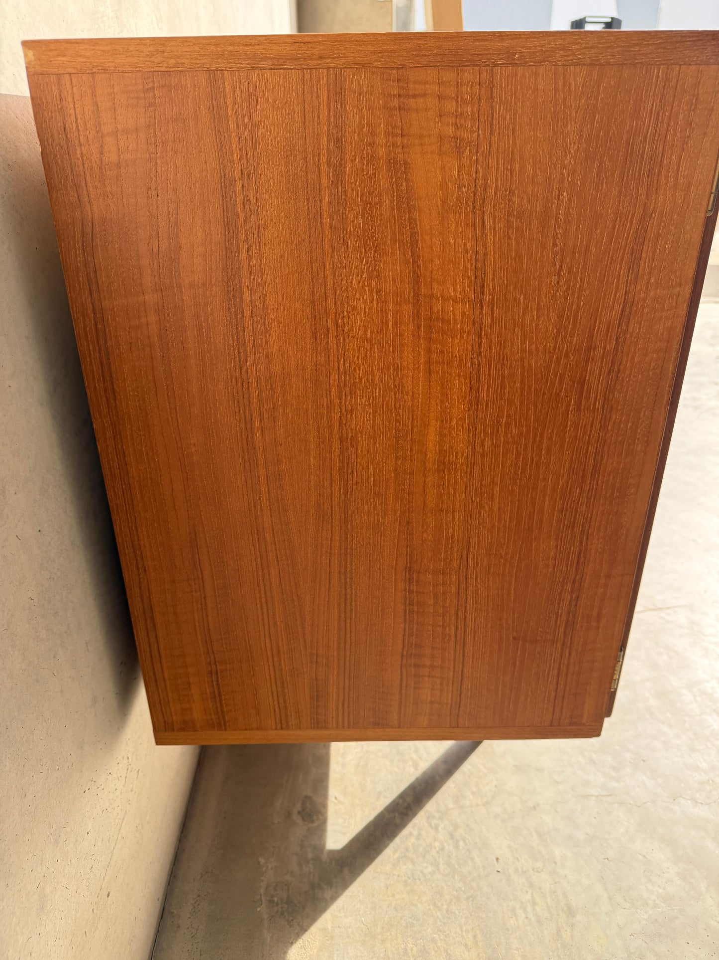 The Iconic Parker Teak 4-Door Sideboard – Restored Mid Century Modern with Tapered Legs (Rediscover) features natural brown wood grain and stands upright against a beige wall, casting a shadow on a light-colored floor.
