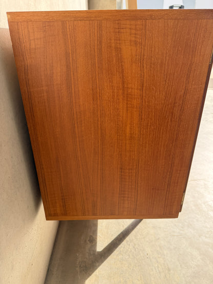 The Iconic Parker Teak 4-Door Sideboard – Restored Mid Century Modern with Tapered Legs (Rediscover) features natural brown wood grain and stands upright against a beige wall, casting a shadow on a light-colored floor.