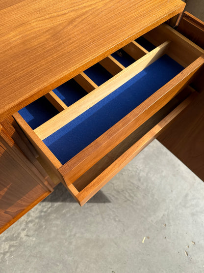 An open drawer of the Iconic Parker Teak 4-Door Sideboard – Restored Mid Century Modern with Tapered Legs reveals blue felt lining and dividers, highlighting Australian craftsmanship, bathed in sunlight on a concrete floor.