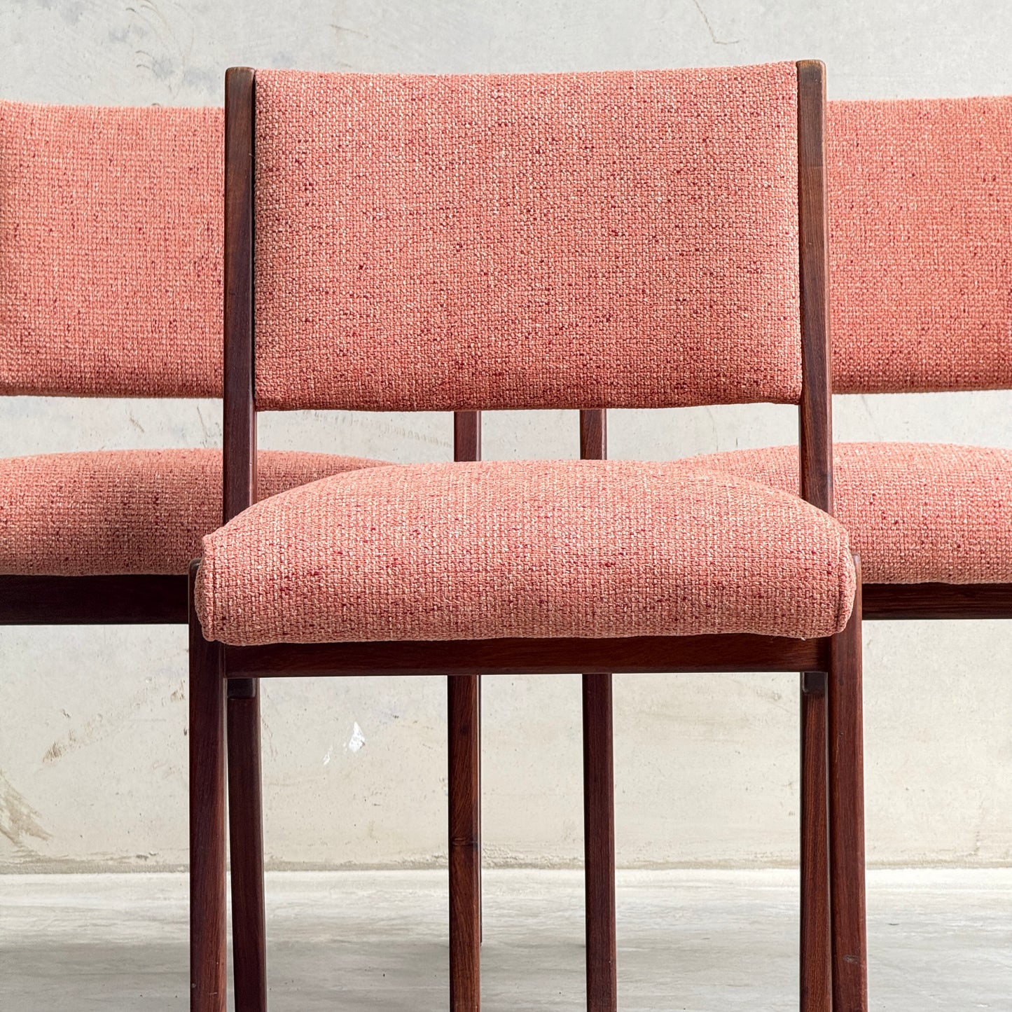 A collectable set of 4 Vintage MCM Jens Risom Chairs is arranged against a light wall and concrete floor, highlighting their Scandinavian design with pink textured fabric on the seats and backs. The focus is on the center chair.