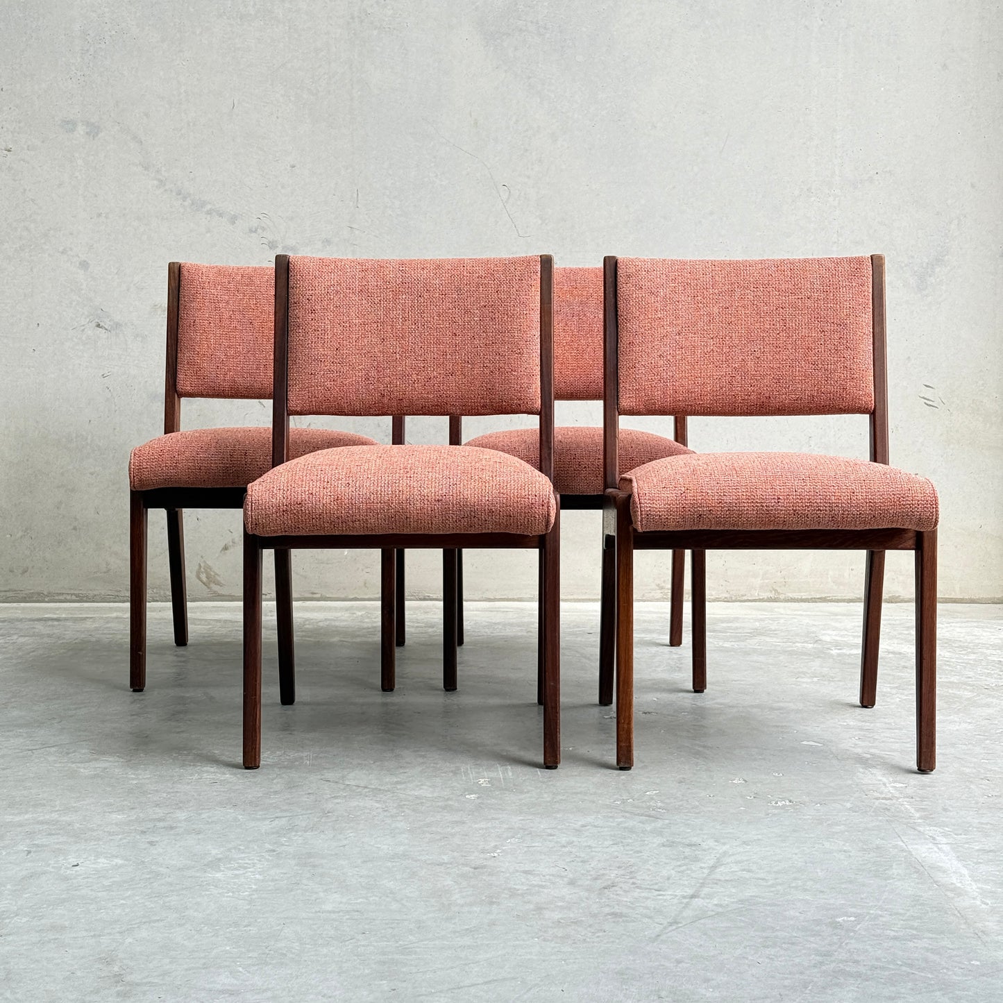 A collectable set of 4 Vintage MCM Jens Risom Chairs with reddish-pink upholstered seats and wooden frames is arranged on a concrete floor, showcasing timeless Scandinavian design against a plain, light gray wall.