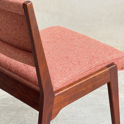A close-up of a Vintage MCM Jens Risom Chair from a collectable set of 4 by Refine, showing its reddish-pink textured upholstery and smooth, angled wooden frame against a plain, light-colored background.