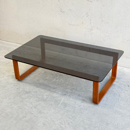The Tessa T4 - Large Coffee Table (Revive), featuring a dark glass top and wooden legs, stands on a light gray concrete floor against a plain, light-colored wall.