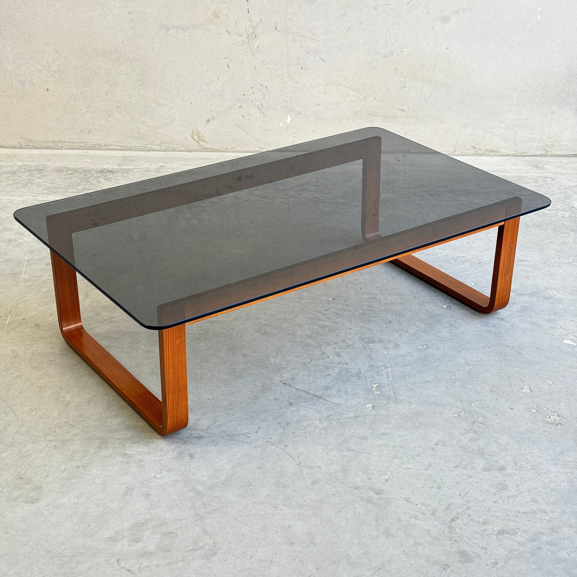The Tessa T4 - Large Coffee Table (Revive) features a glass top and rounded wooden legs, placed on a smooth light gray concrete floor with a plain, light-colored wall in the background.