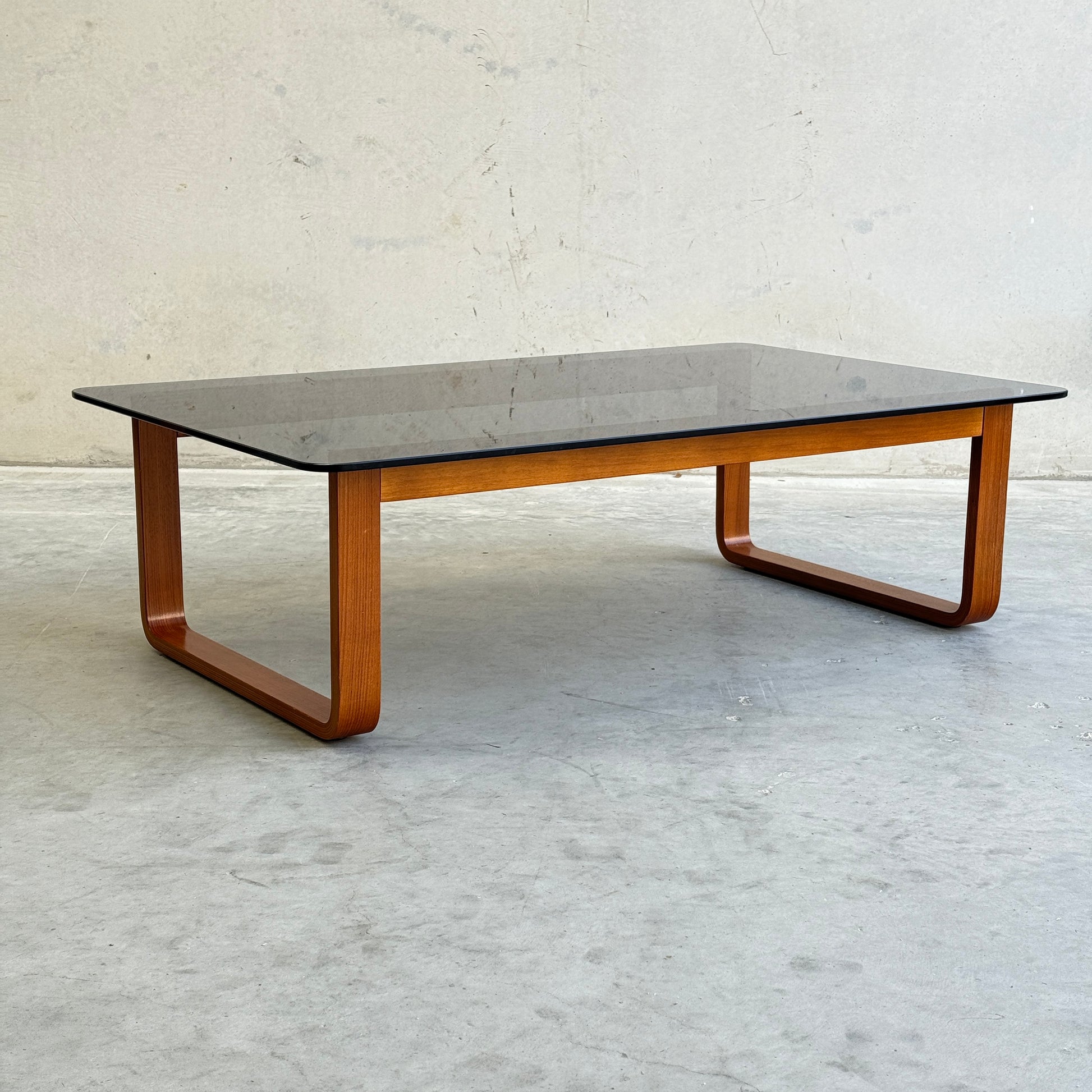 The Tessa T4 - Large Coffee Table (Revive) features a minimalist design with a rectangular glass top and curved wooden legs, set on a smooth light gray floor against a plain textured wall.