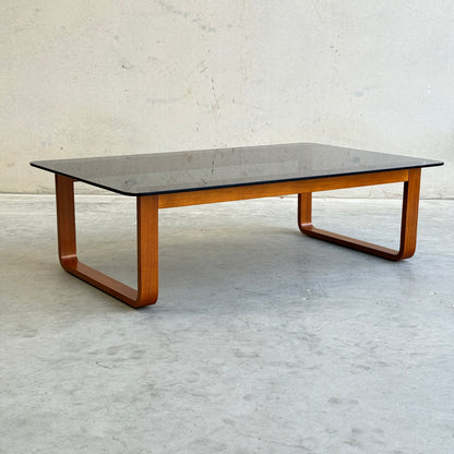 The Tessa T4 - Large Coffee Table (Revive) features a minimalist design with a rectangular glass top and curved wooden legs, set on a smooth light gray floor against a plain textured wall.