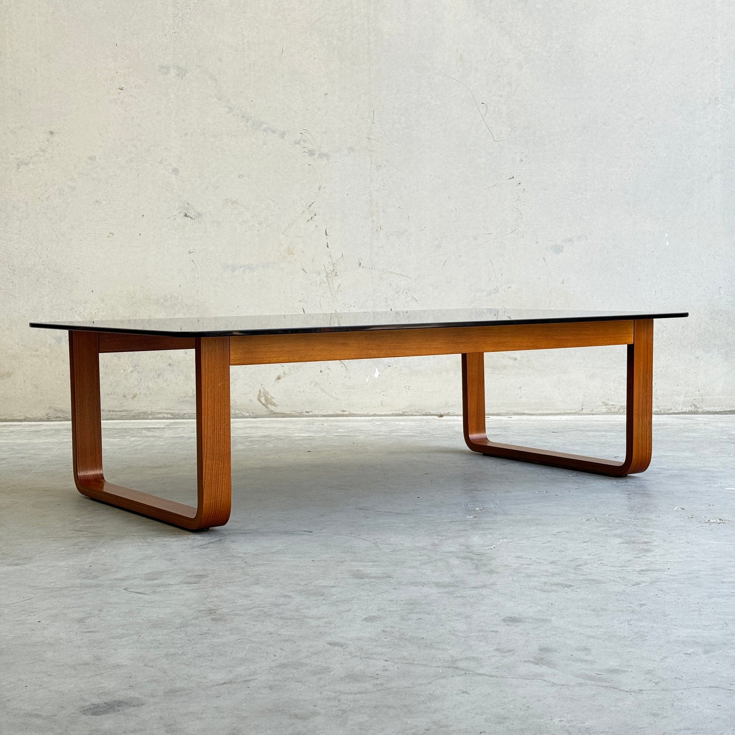 The Tessa T4 - Large Coffee Table (Revive) features a sleek rectangular black top and curved wooden legs, combining modern minimalist style with the elegance of a wood frame—perfect against any light wall and concrete floor.