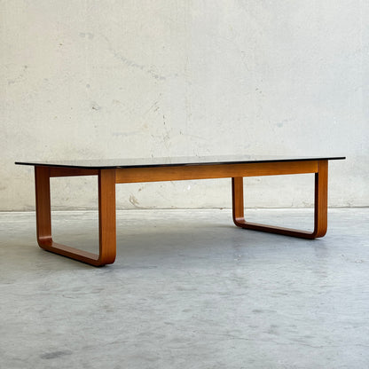 The Tessa T4 - Large Coffee Table (Revive) features a sleek rectangular black top and curved wooden legs, combining modern minimalist style with the elegance of a wood frame—perfect against any light wall and concrete floor.