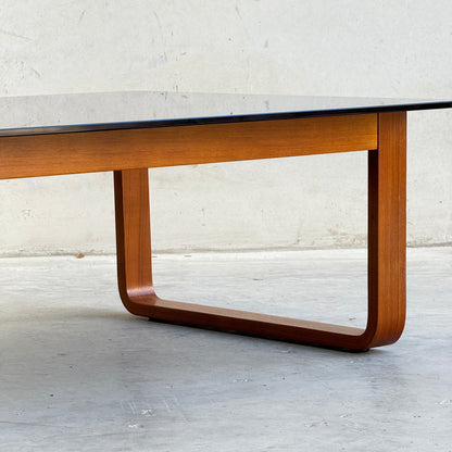 A close-up of the Tessa T4 Large Coffee Table (Revive), showcasing its rectangular glass top and curved wooden legs, set against a plain, light-colored wall and floor.