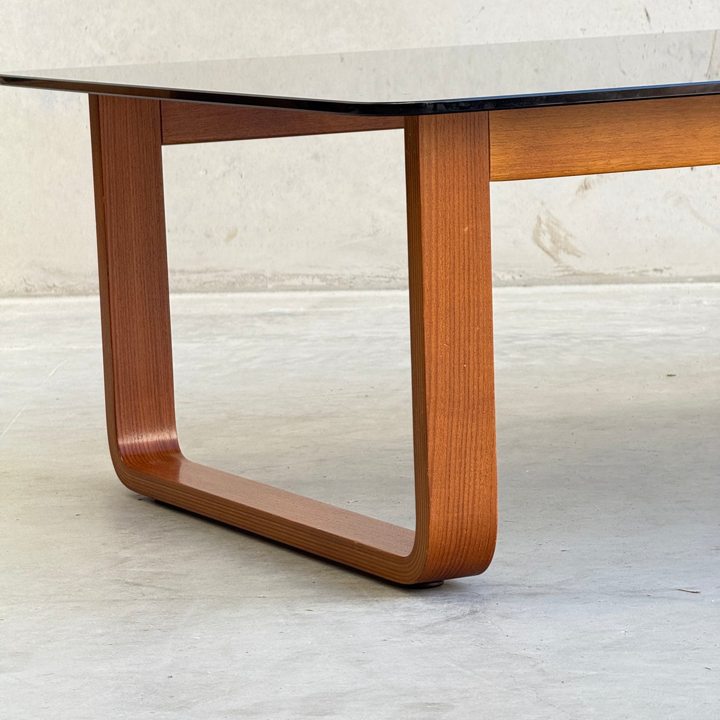 A close-up of the Tessa T4 - Large Coffee Table (Revive), featuring a rectangular black glass top and curved wooden leg, displayed on a light surface. Its design is modern, minimalist, and contemporary.