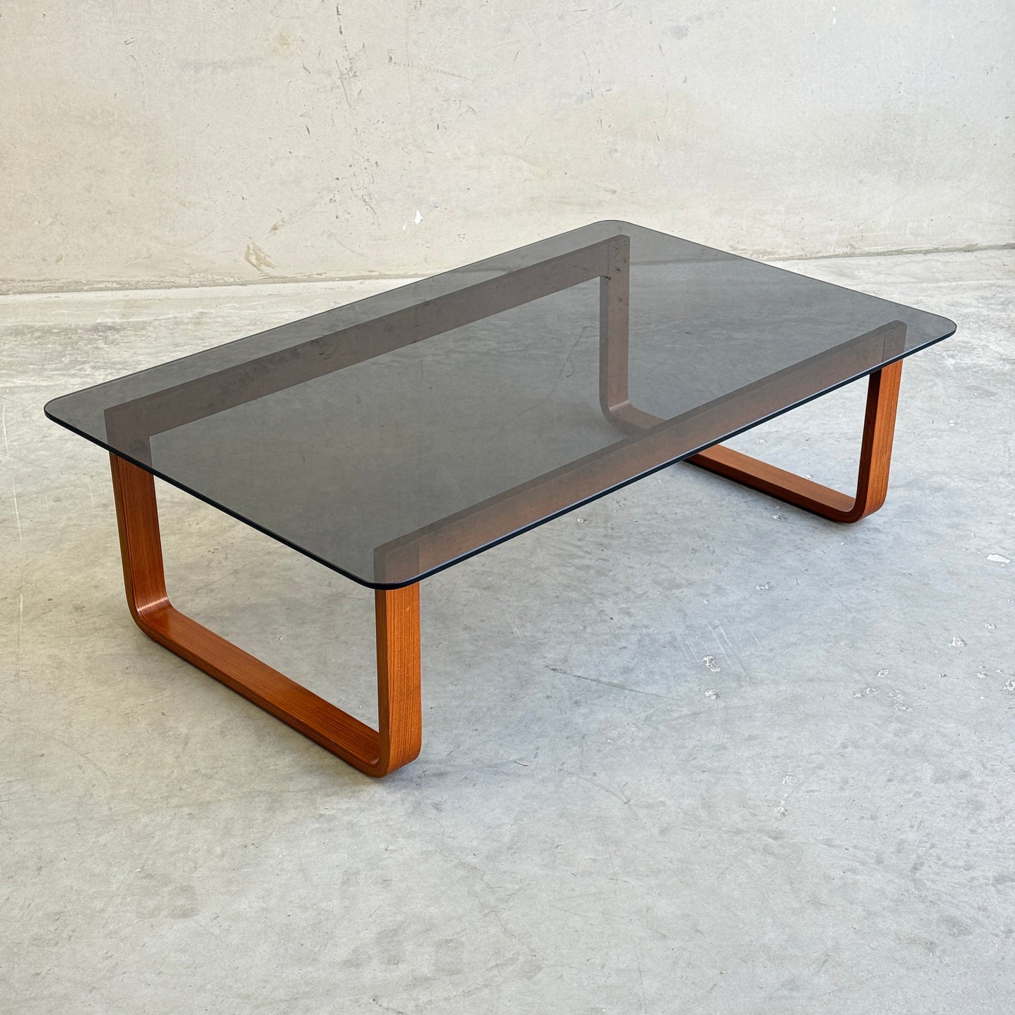 The Tessa T4 - Large Coffee Table (Revive), featuring a modern minimalist glass top, rounded edges, and wooden sled-style legs, stands on a concrete floor against a plain light-colored wall.