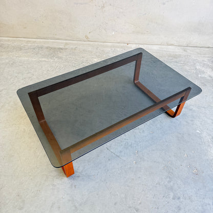 The Tessa T4 Large Coffee Table (Revive), featuring a dark tinted glass top and wooden legs, stands on a light gray concrete floor against a plain wall.