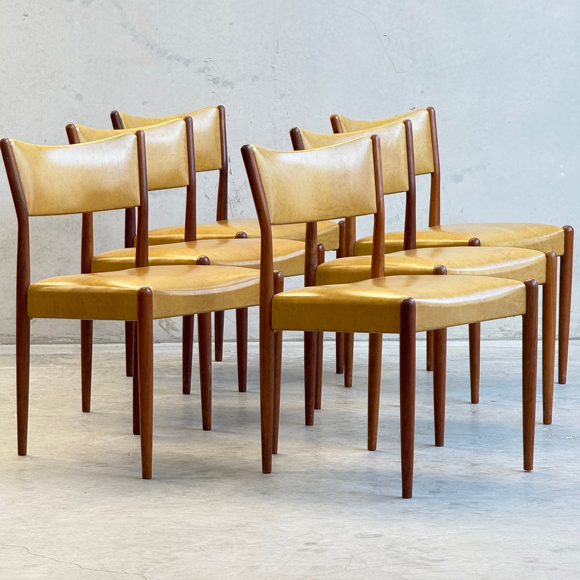A set of six Parker Number 97 Dining Chairs in original mustard vinyl is arranged in two rows on a light floor, displayed against a plain gray wall.