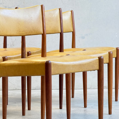 Six Parker Number 97 dining chairs with original mustard vinyl upholstery are arranged in a staggered row against a light gray wall.