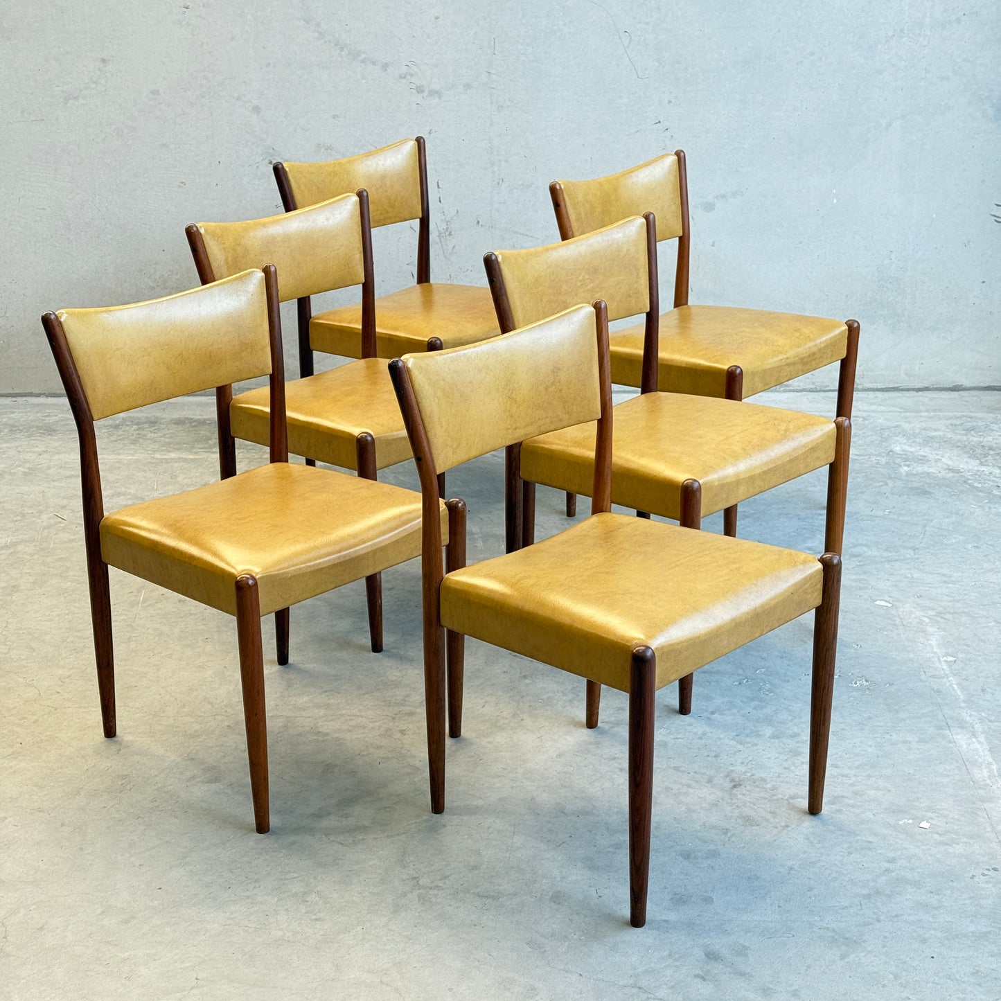 Six Parker Number 97 Dining Chairs, featuring original mustard vinyl seats and backs with dark wooden legs, are arranged in two rows on a gray concrete floor against a plain gray wall.