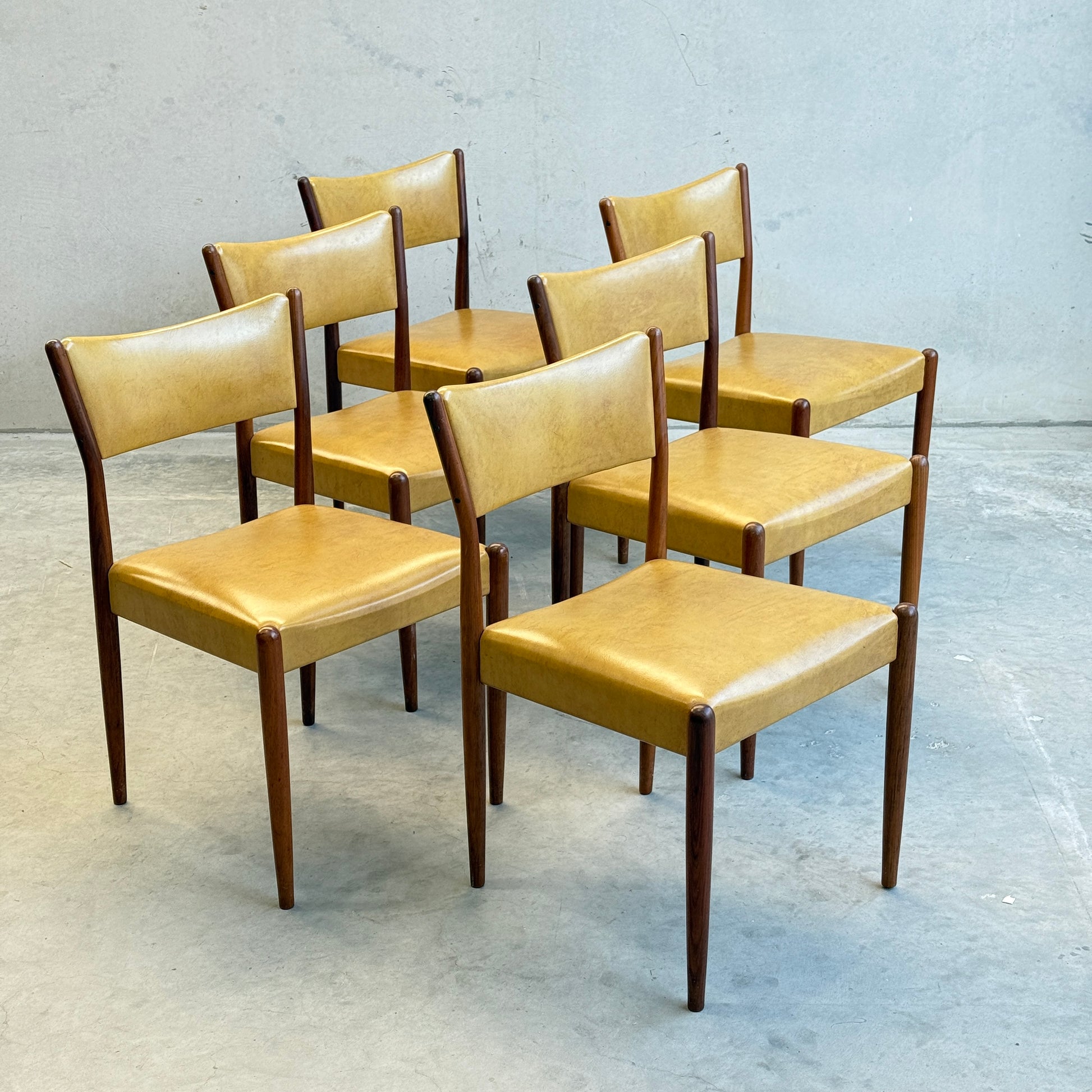 Six Parker Number 97 Dining Chairs, featuring original mustard vinyl seats and backs with dark wooden legs, are arranged in two rows on a gray concrete floor against a plain gray wall.