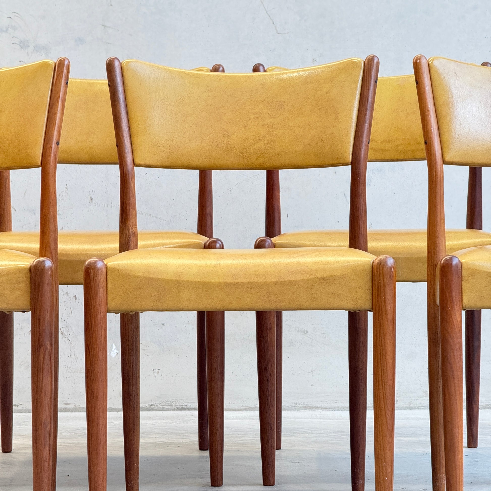 A set of six Parker Number 97 Dining Chairs with original mustard vinyl upholstery, arranged together against a light grey wall, embodying classic mid-century modern style.
