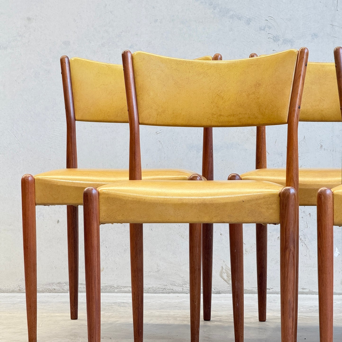 Six Parker Number 97 dining chairs in original mustard vinyl are arranged in a staggered row against a light gray wall, highlighting their classic mid-century modern style.