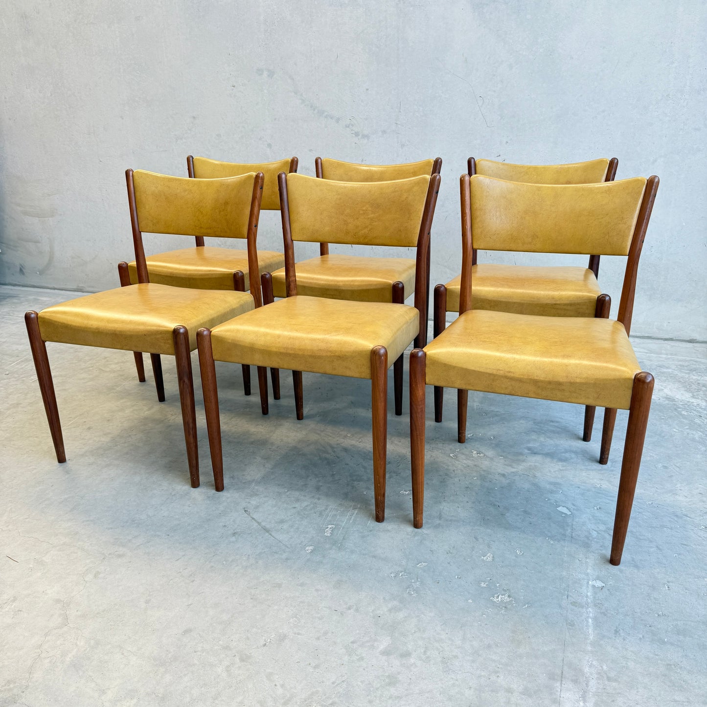 A set of six Parker Number 97 Dining Chairs in original mustard vinyl is arranged in two rows on a concrete floor against a light wall, highlighting their classic mid-century modern style.