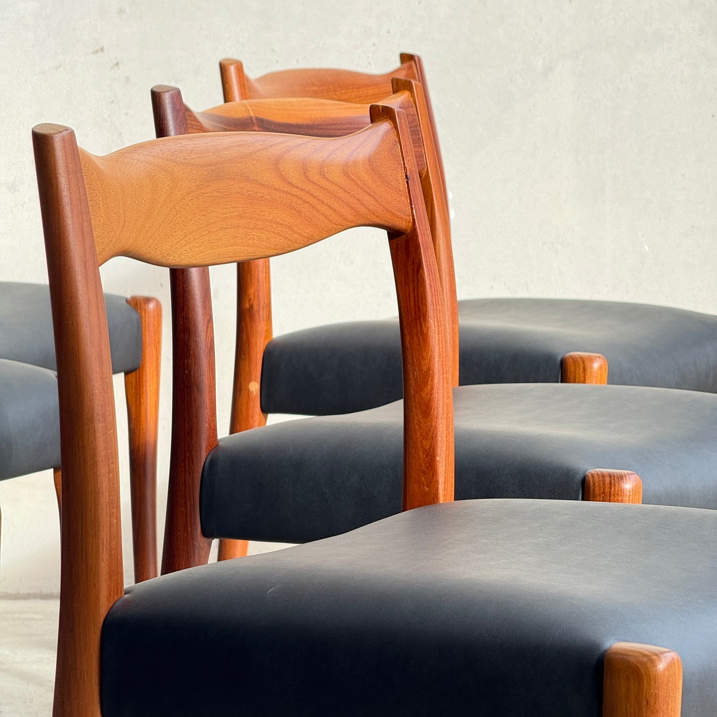 A fully restored set of 6 Fler 64 dining chairs with sculpted backs and liquorice vinyl seats is arranged in a row against a plain, light wall, where the warm wood contrasts stylishly with the dark upholstery.