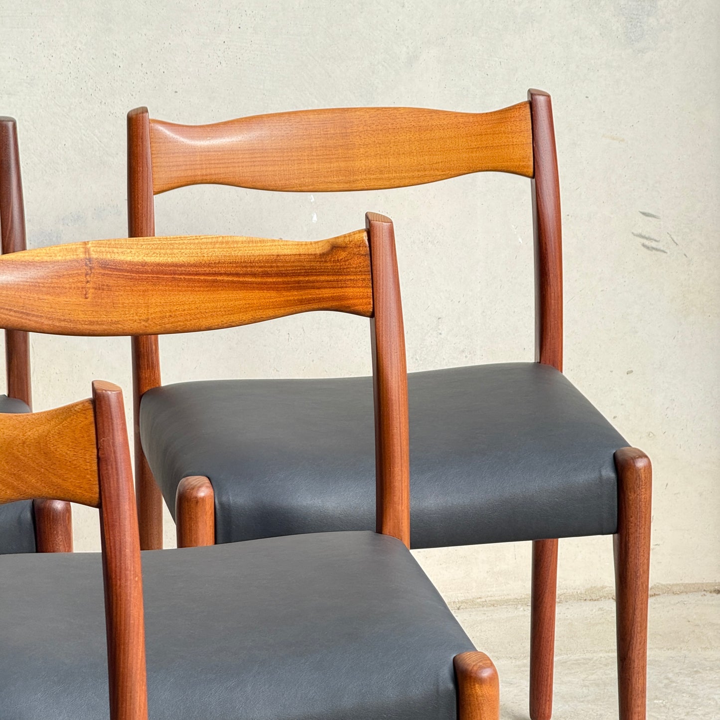 Six fully restored mid-century Fler 64 blackwood chairs from the Parker 91 Oval Teak Dining Suite, featuring smooth black seats and warm wood tones, are arranged in front of a light-colored wall.