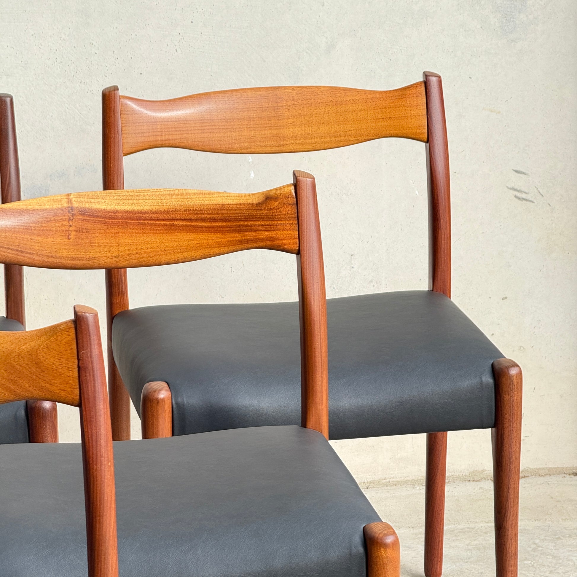 Six fully restored mid-century Fler 64 blackwood chairs from the Parker 91 Oval Teak Dining Suite, featuring smooth black seats and warm wood tones, are arranged in front of a light-colored wall.