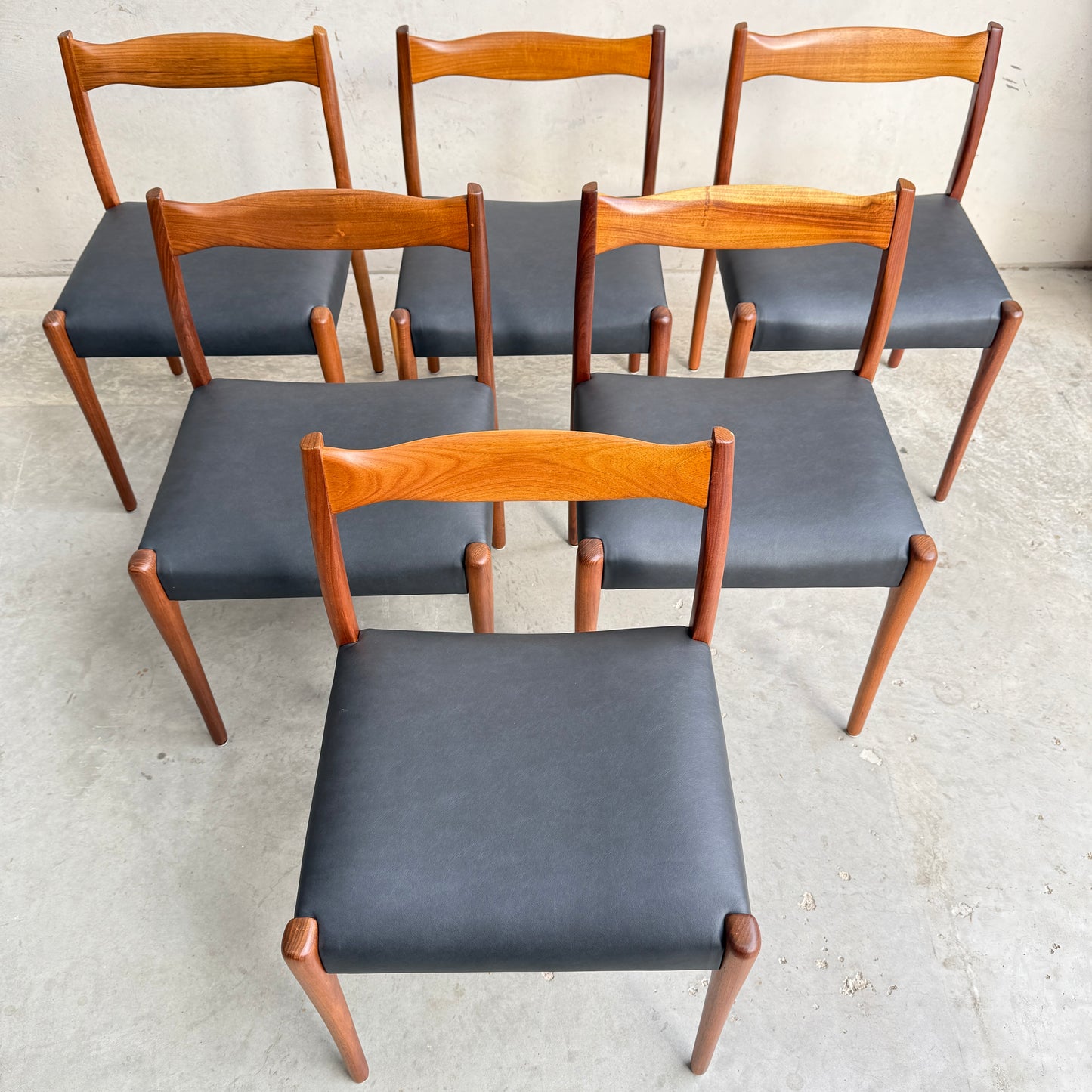 A fully restored set of 6 Fler 64 dining chairs in liquorice vinyl (Rediscover) is arranged on a concrete floor in two uneven rows, their mid-century modern elegance standing out against a plain light wall.