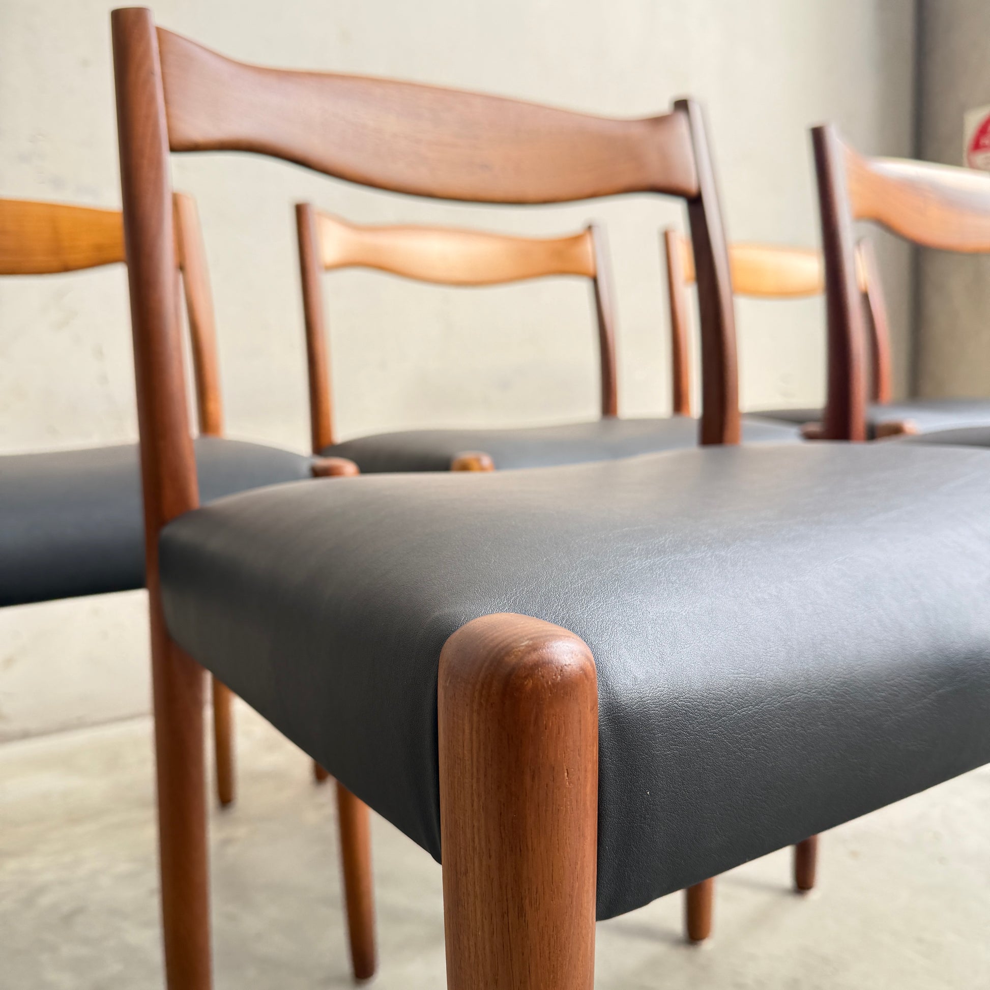 A close-up of a fully restored set of 6 Fler 64 dining chairs with curved wooden backs and liquorice vinyl seats, arranged in a row against a plain, light-colored wall.