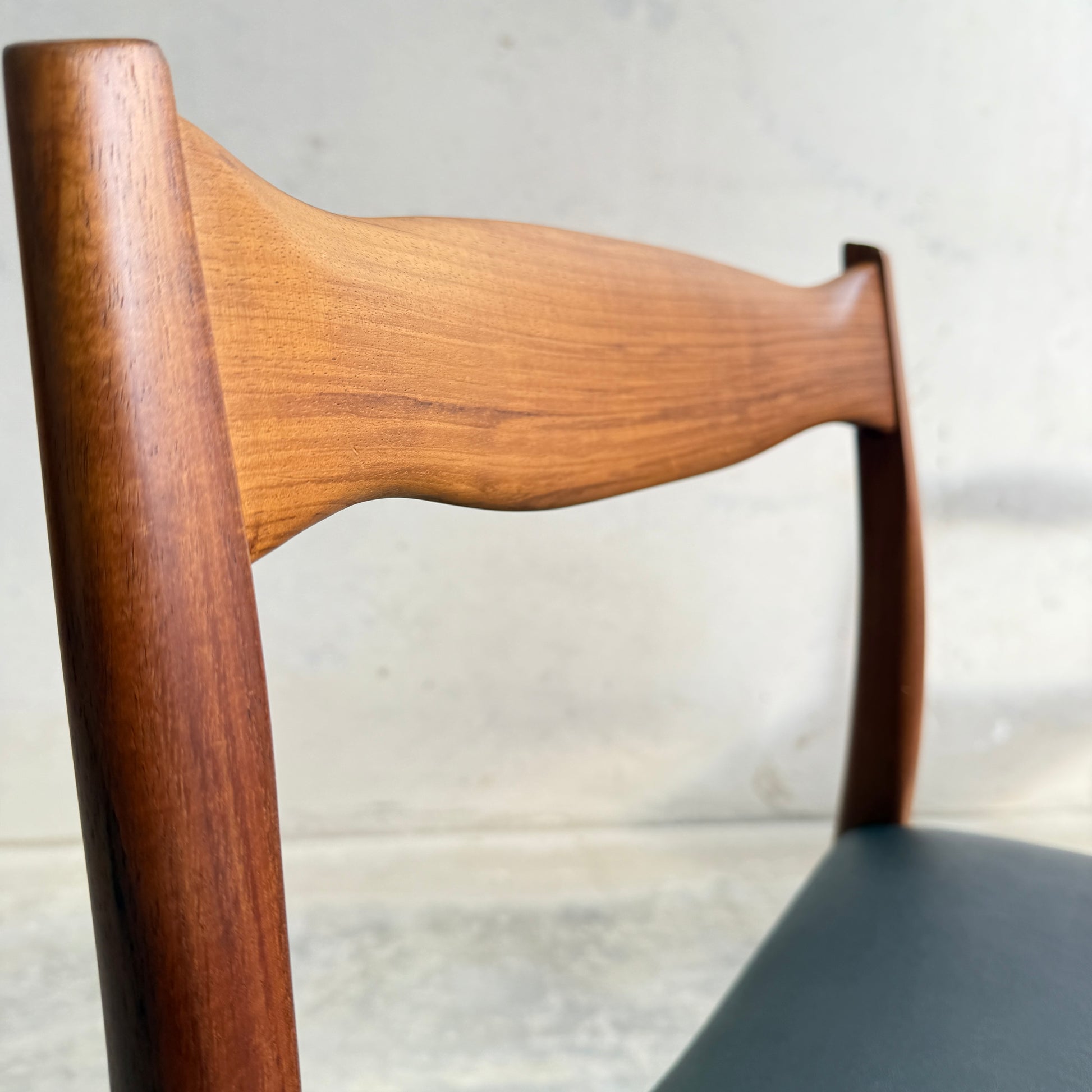 Close-up of a Fully Restored Fler 64 dining chair backrest in Liquorice Vinyl, showcasing a smooth curved design with matte finish. The dark seat contrasts with the light, textured wall, reflecting mid-century modern style. Set includes 6 chairs.