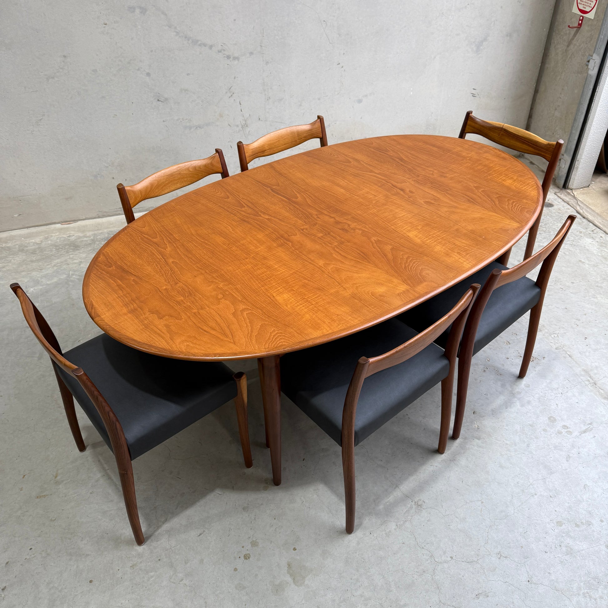 A fully restored Mid-Century Parker 91 oval teak dining suite with six Fler 64 blackwood chairs (Rediscover) is showcased on a light gray concrete floor against a plain, light-colored wall.