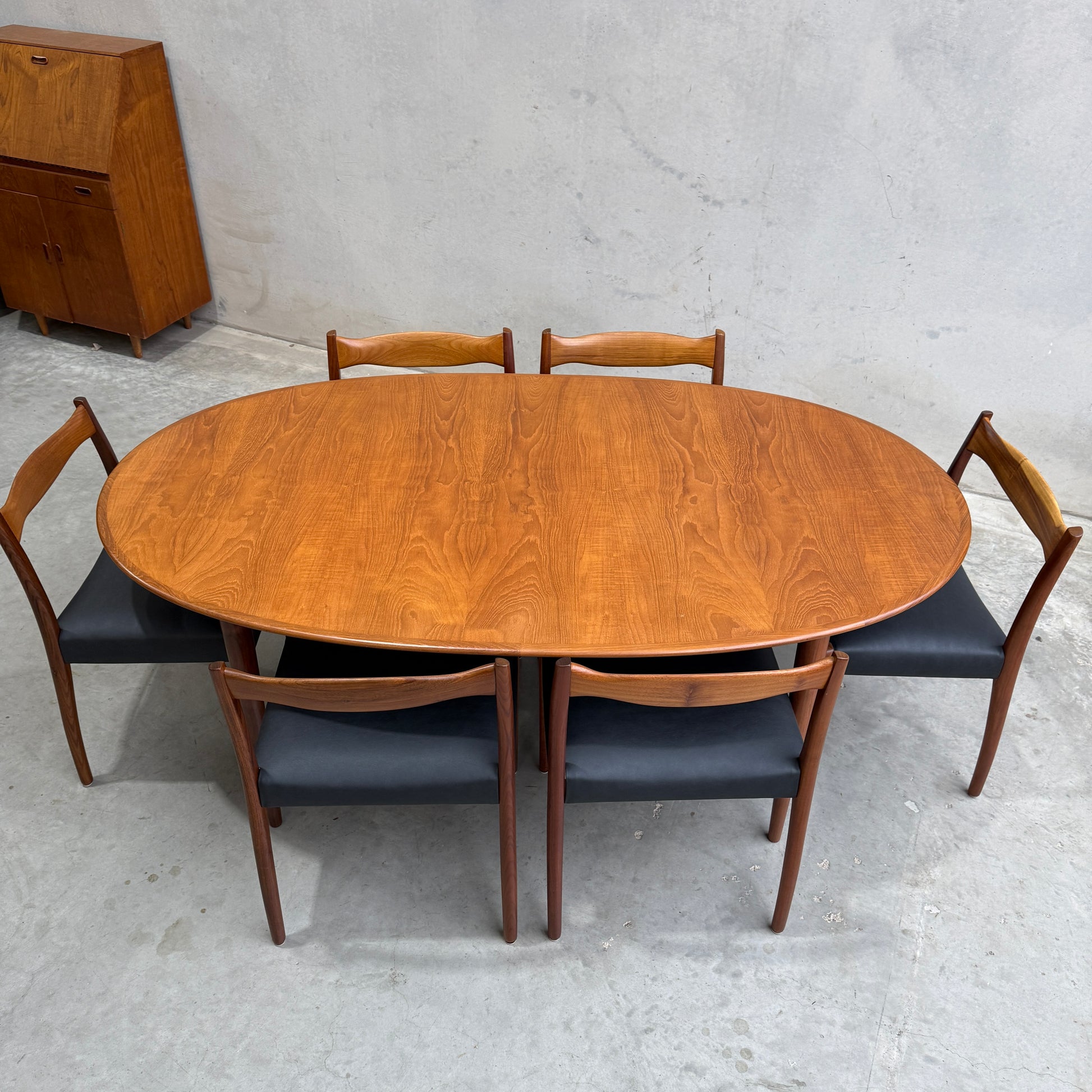 Fully Restored Mid-Century Parker 91 Oval Teak Dining Suite with 6 Fler 64 Blackwood Chairs, featuring dark cushions. Set on a light gray concrete floor with a wooden cabinet in the background.