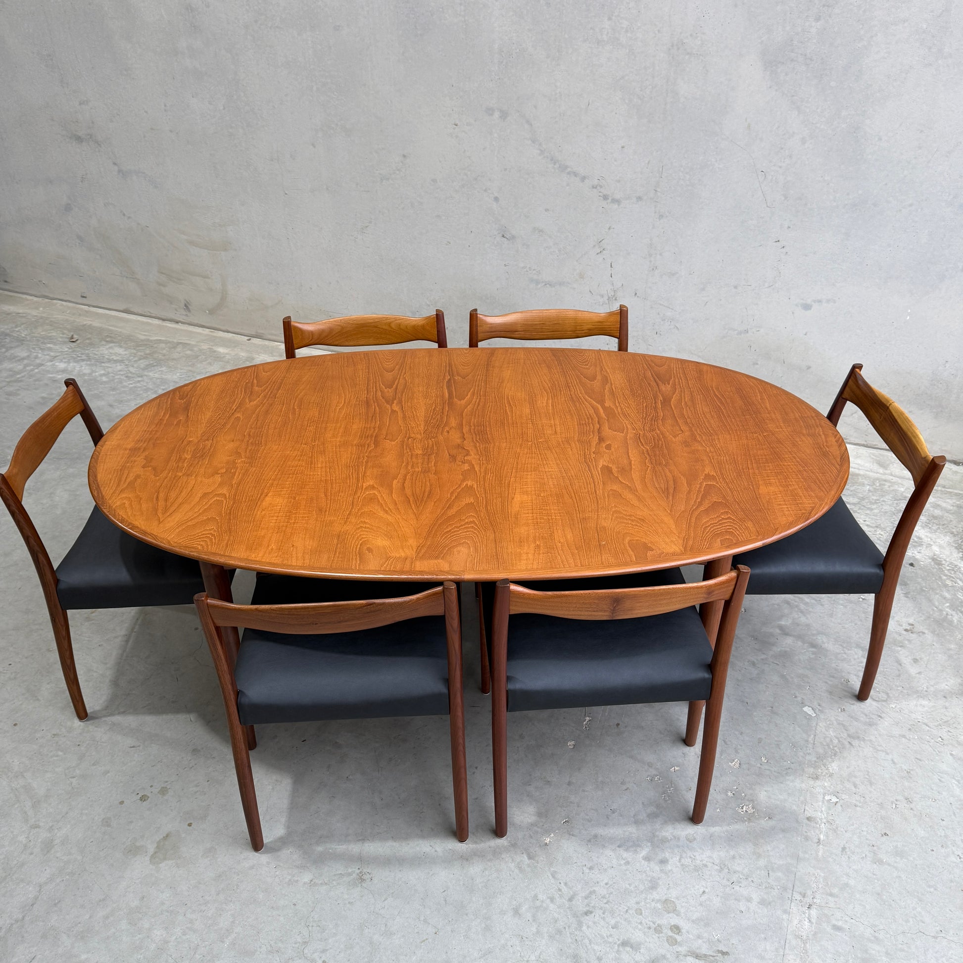 A Fully Restored Mid-Century Parker 91 Oval Teak Dining Suite with 6 Fler 64 Blackwood Chairs (Rediscover) sits on a gray concrete floor against a light gray wall.