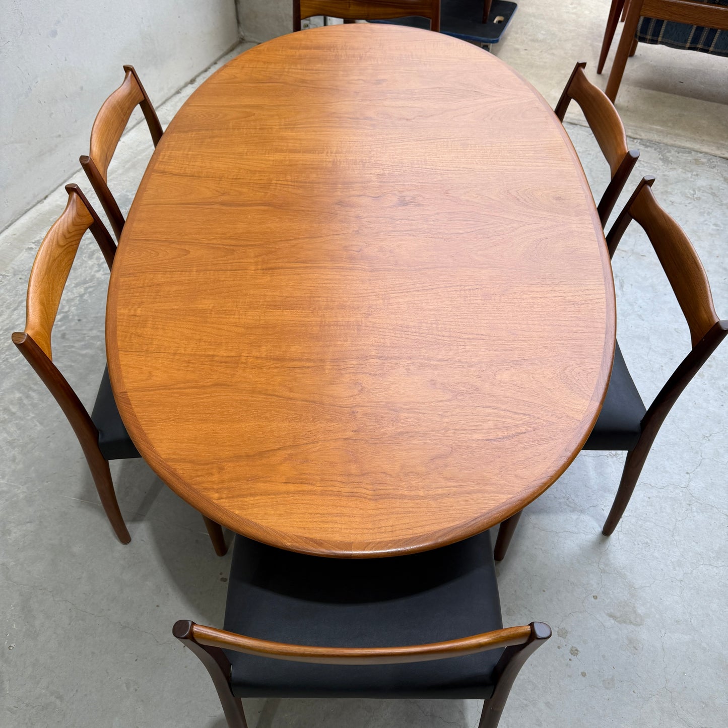 A fully restored mid-century Parker 91 oval teak dining suite with six Fler 64 blackwood chairs is arranged on a light concrete floor, showcasing its beautiful natural wood grain finish.