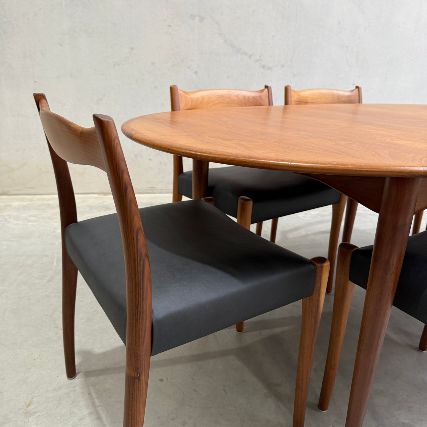 A close-up of a fully restored mid-century Parker 91 oval teak dining suite with six Fler 64 blackwood chairs, set on a plain concrete floor against a light-colored wall.