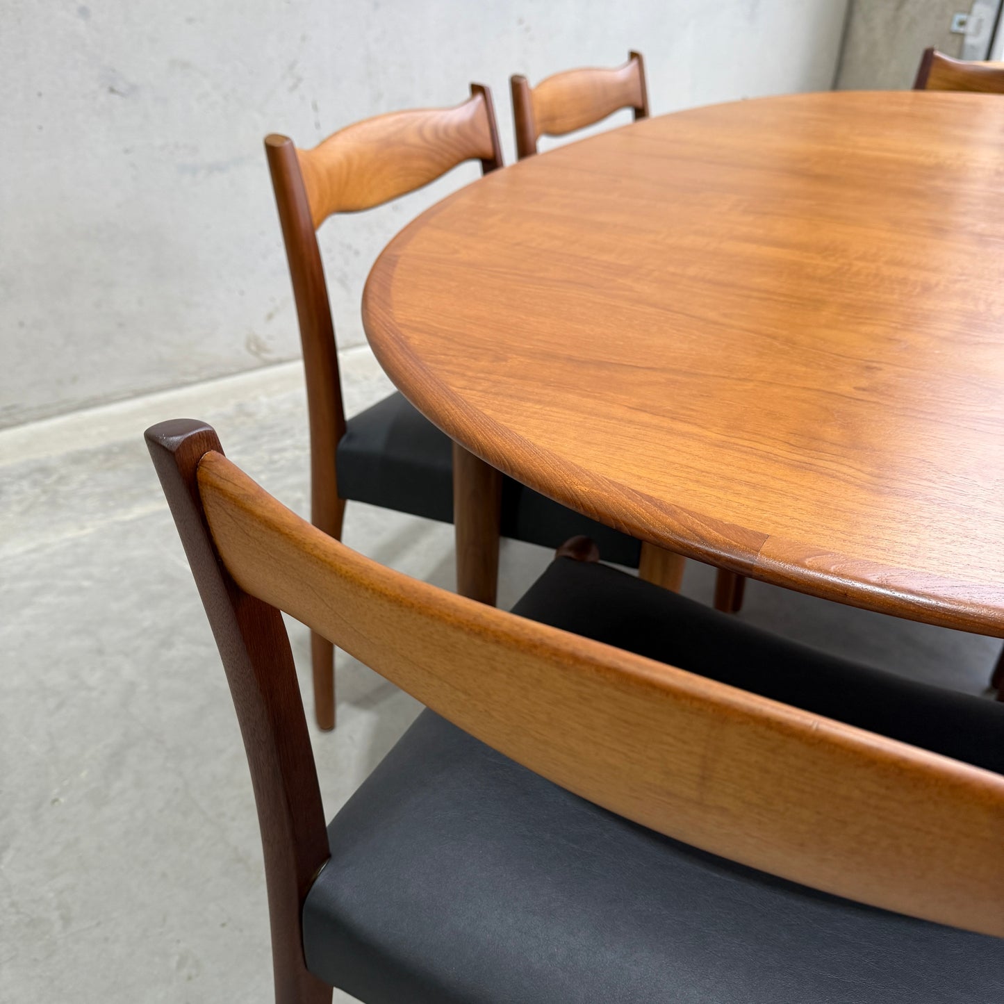 A close-up of the Fully Restored Mid-Century Parker 91 Oval Teak Dining Suite with 6 Fler 64 Blackwood Chairs (Rediscover), showcasing the set’s sculpted backs and black cushions against a simple concrete wall and floor.