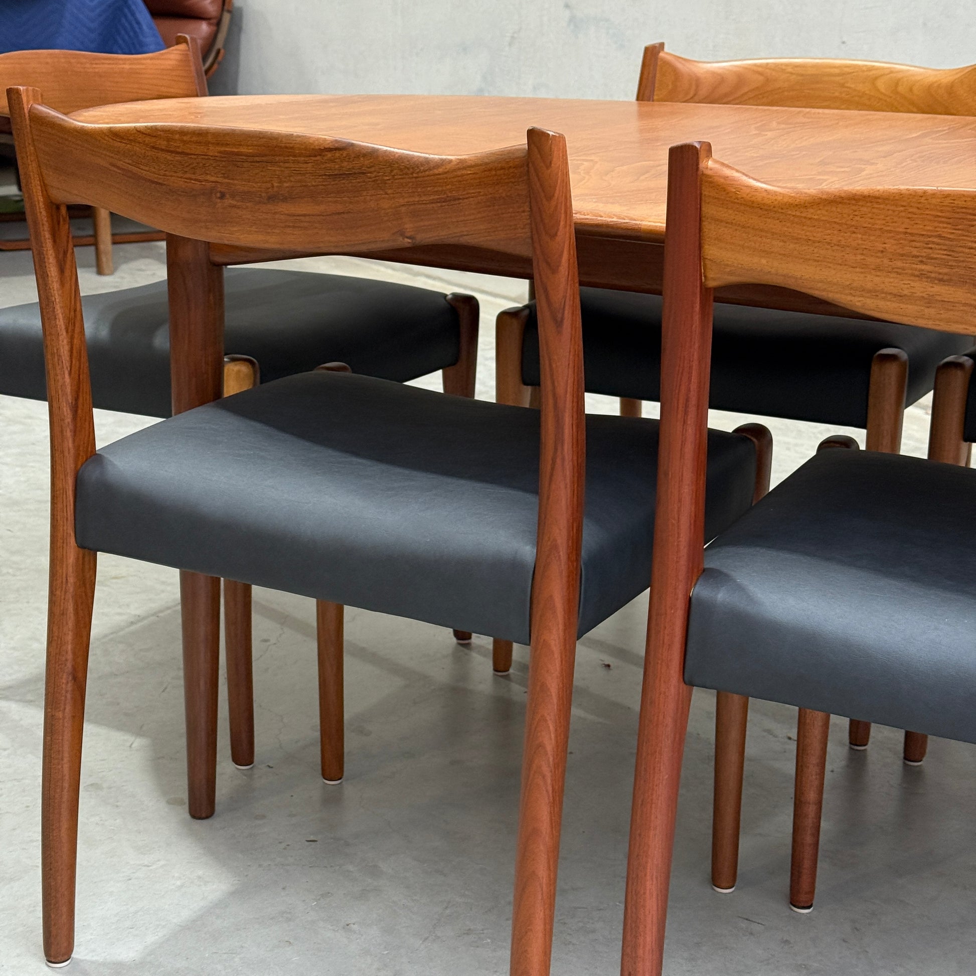 A close-up of the Fully Restored Mid-Century Parker 91 Oval Teak Dining Suite with 6 Fler 64 Blackwood Chairs (Rediscover) featuring curved backs and black upholstered seats on a concrete floor.