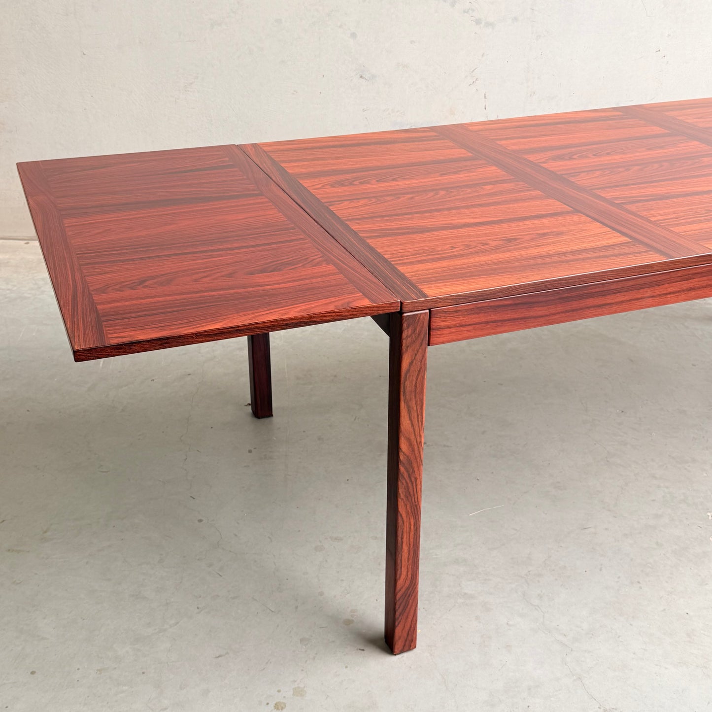 A 1970s Danish Modern Rosewood Dropleaf Dining Table by Vejle Stole og Mobelfabrik (Model 166, 1975), featuring a rich reddish-brown grain and sturdy rectangular legs, stands against a light wall and concrete floor.