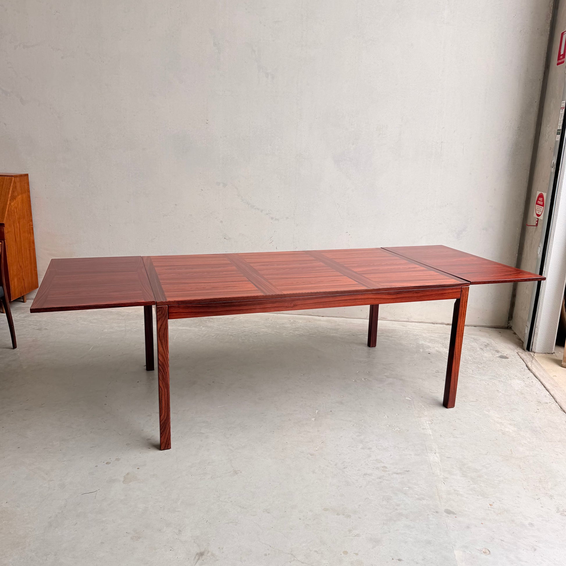 A 1970s Danish Modern Rosewood Dropleaf Dining Table by Vejle Stole og Mobelfabrik (Model 166, 1975), with four straight legs and extended leaves, stands in a minimalist room with a concrete floor and plain light wall.