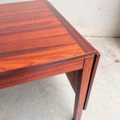 A close-up of the 1970s Danish Modern Rosewood Dropleaf Dining Table by Vejle Stole og Mobelfabrik (Model 166, 1975) displays its rich rosewood grain and foldable extension leaf, set on a light concrete floor.