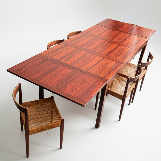 A 1970s Danish Modern Rosewood Dropleaf Dining Table by Vejle Stole og Mobelfabrik (Model 166, 1975) with a glossy surface is surrounded by six woven-seat chairs in a minimalist room with concrete flooring and a blank wall.