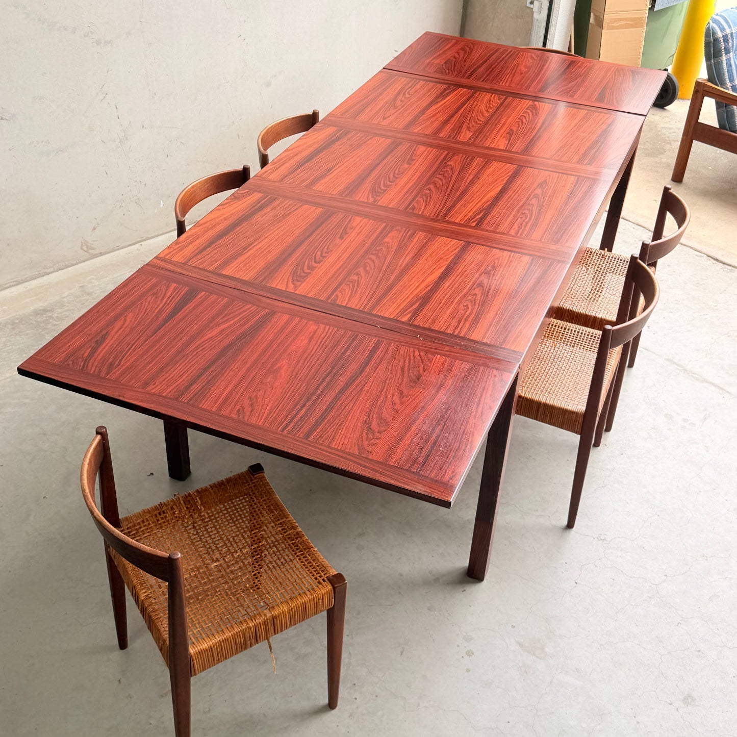 A 1970s Danish Modern Rosewood Dropleaf Dining Table by Vejle Stole og Mobelfabrik (Model 166, 1975), with a rich striped grain, stands surrounded by cane-seat chairs in a minimalist mid-century room with a light-toned floor.