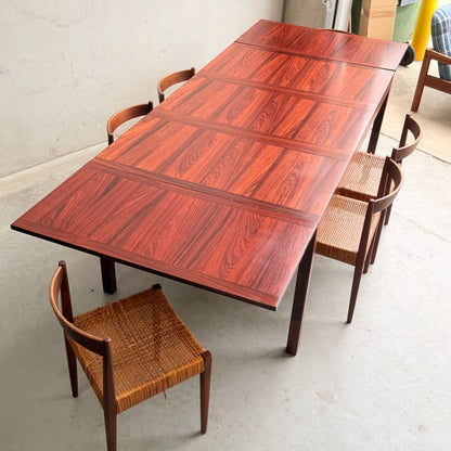 A 1970s Danish Modern Rosewood Dropleaf Dining Table by Vejle Stole og Mobelfabrik (Model 166, 1975), with a rich striped grain, stands surrounded by cane-seat chairs in a minimalist mid-century room with a light-toned floor.