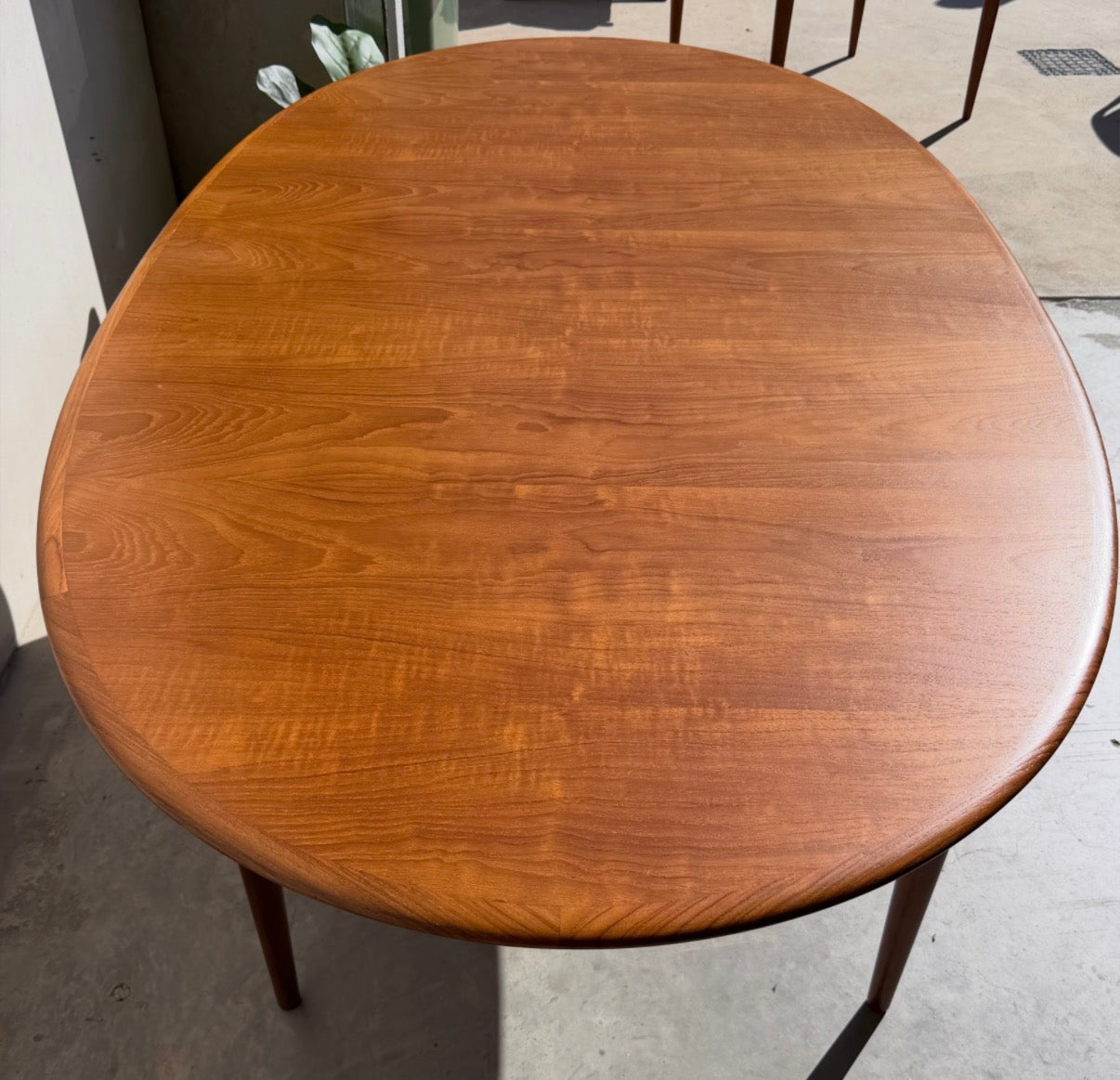 A fully restored mid-century Parker 91 oval teak dining suite with 6 Fler 64 blackwood chairs (Rediscover) stands on a light floor in a sunlit room, its polished surface and visible grain offering stylish elegance.